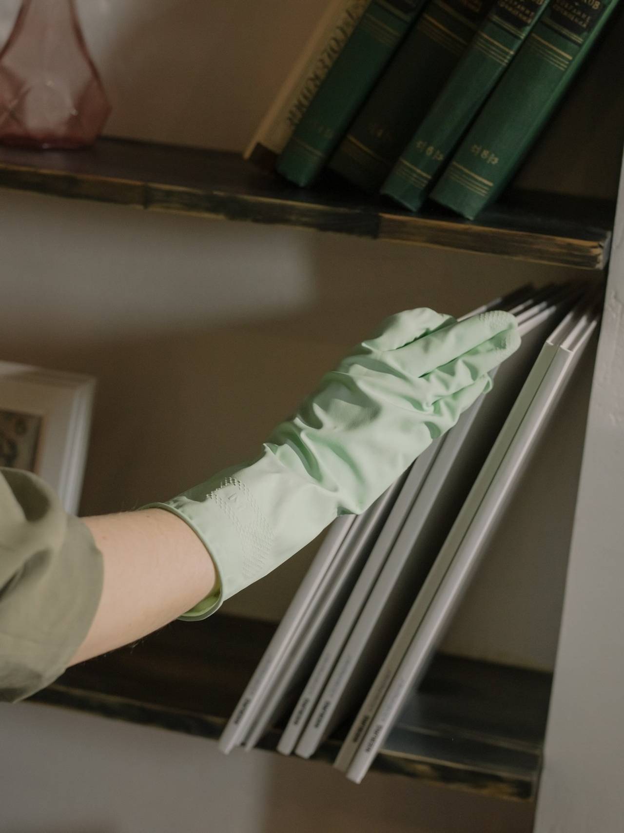 Mão com luva verde retirando alguns livros brancos e finos de uma prateleira. Na prateleira de cima alguns livros verdes.