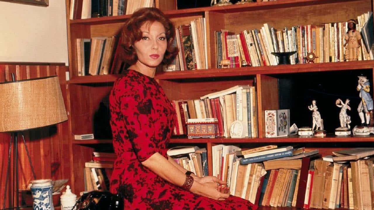 Foto de Clarice Lispector com um vestido vermelho em frente a uma estante de livros e ao lado de um abajur